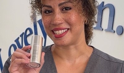 A woman with a medium skin tone and curly brown and blonde hair smiles while holding Dermalogica Smart Density Undereye Serum.