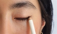 A woman with a light skin tone and dark brown hair presses a bronze eyeliner pencil to her eyelid.