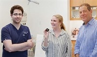 Three people stand candidly in a doctor's office smiling at one another or the camera.