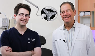 Two dermatologists, one with fair skin and dark hair smiles while crossing his arms next to one with light brown hair wearing a white jacket.