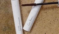 Two mascaras lay next to one another over a granite countertop with a mascara brush laid over top of them.