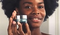 A black woman smiling softly and holding a jar of ELEMIS Eye Revive Masks in front of her cheek.