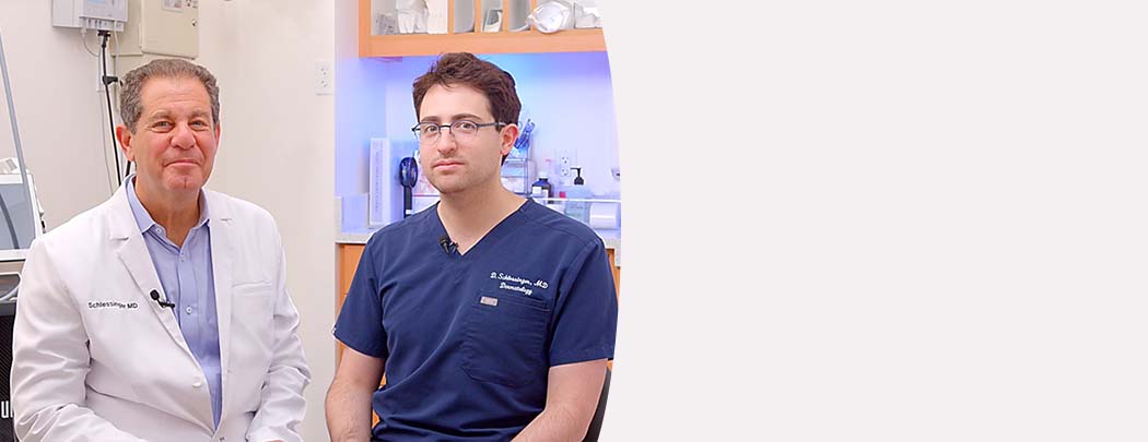 Two dermatologists, one in a white jacket and one in scrubs, sit next to one another in a patient room smiling.
