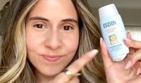 A woman with a medium skin tone and dark brown hair smiles as she points to a blue bottle of ISDIN Fusion Water Magic Broad Spectrum SPF 40 sunscreen.