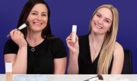 A woman with dark brown hair and light skin holds a makeup brush to her cheek while a blonde woman with fair skin holds a bottle of foundation.