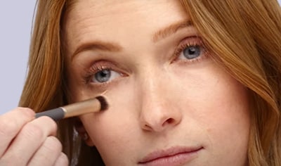 A woman with fair skin and red hair uses a makeup brush to apply Jane Iredale Circle\Delete Concealer beneath her eyes.