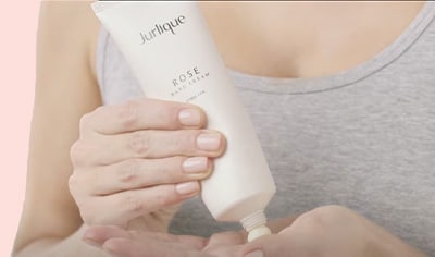 A woman with fair skin, nude colored nails and a grey tank top dispenses Jurlique Rose Hand Cream into her hand.