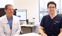 Two dermatologists, one with fair skin and dark hair wearing a dark blue shirt and one with light hair wearing a white jacket, smile at the camera.