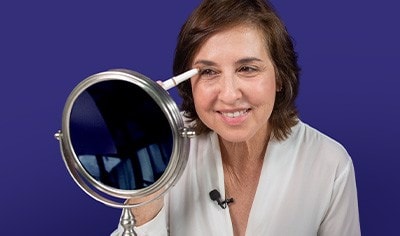 A woman applying an eyeshadow stick to her upper eyelid and smiling in a mirror.