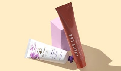 Two tubes of Holifrog and Indie Lee cleansing balms propped up against a pink wooden cube on a beige background.