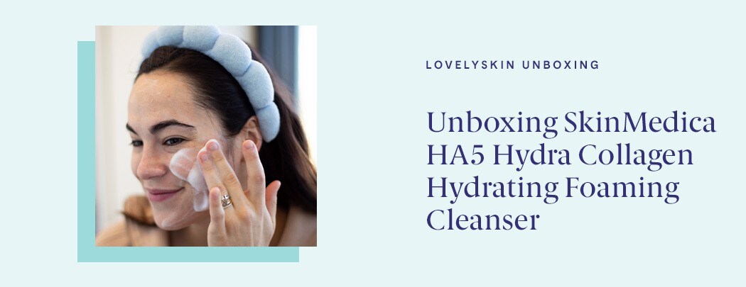 A woman with a medium skin tone and dark brown hair wearing a light blue headband applies a foaming cleanser to her face with a soft smile.