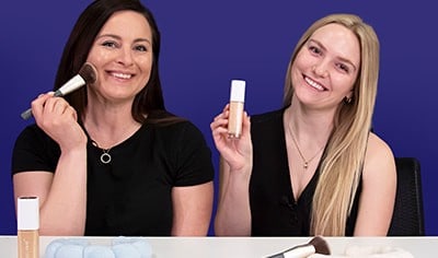 A woman with dark brown hair and light skin holds a makeup brush to her cheek while a blonde woman with fair skin holds a bottle of foundation.