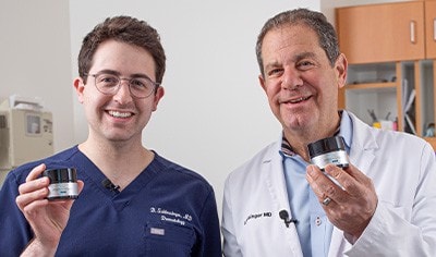 Two dermatologists, one in his 60s and one in his 30s, stand next to one another smiling as they hold SkinCeuticals P-TIOX Micro-Peptide Cream.