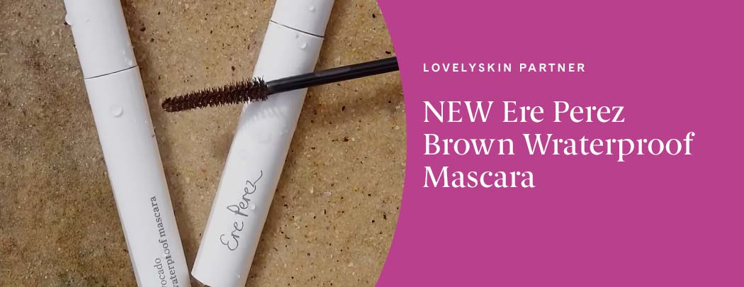 Two mascaras lay next to one another over a granite countertop with a mascara brush laid over top of them.