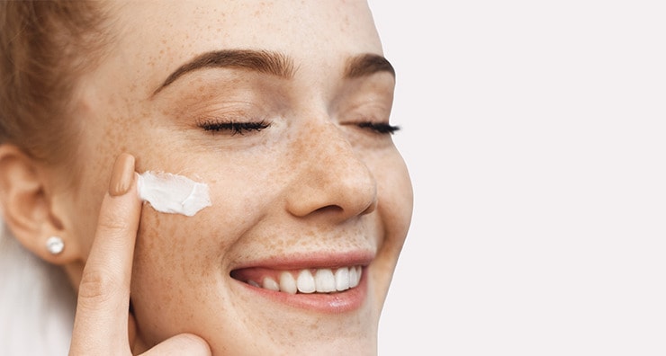 A fair-skinned model with freckles with her eyes closed applying moisturizer to her cheek.