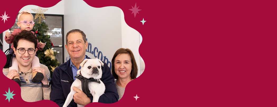 A man in his 30s holds an infant girl on his shoulders while his father and mother smile next to them holding their white pug dog.