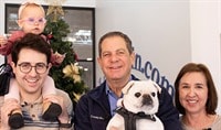 A man in his 30s holds an infant girl on his shoulders while his father and mother smile next to them holding their white pug dog.