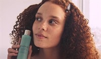 A woman with freckles and red curly hair looking into the distance and holding a bottle of ELEMIS Marine Moisture Essence toner.