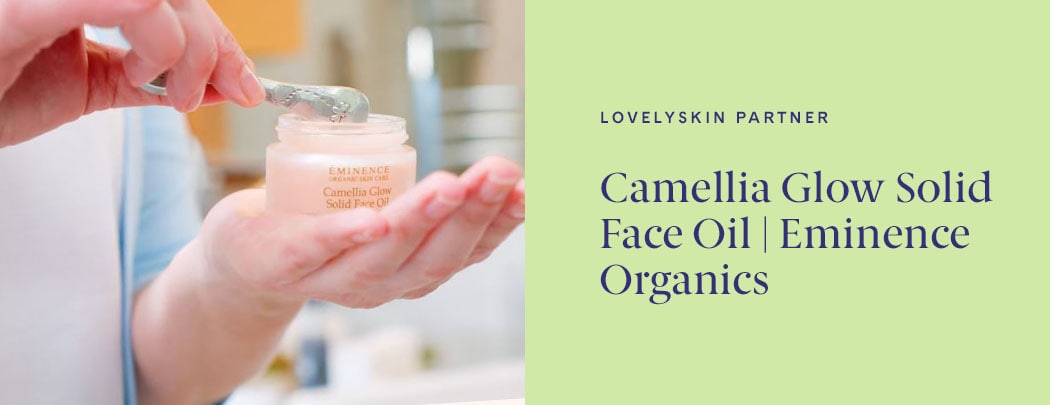 One hand holding a jar of Eminence Organics Camellia Glow Solid Face Oil while the other uses a metal spoon to scoop the product