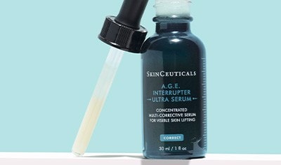 A brown glass bottle of SkinCeuticals A.G.E. Interrupter Ultra Serum with the dropper lid unscrewed and placed next to it on a light blue background.