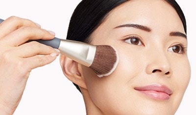 A woman of asian descent with dark brown hair and brown eyes smiles while pressing a makeup brush onto her cheek.