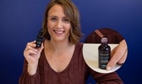 A woman with brown hair holding a bottle of SkinCeuticals HAI Multi-Glycan Multi-dimensional Volumizing Serum and smiling.