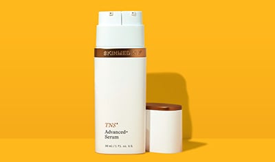 A white bottle with bronze details that reads "TNS Advanced+ Serum" stands on a yellow background.