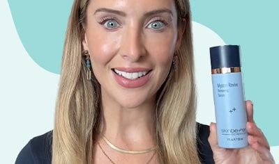 A woman with fair skin and blonde hair holds a bottle of skinbetter science Mystro Revive Renewing Serum while smiling.