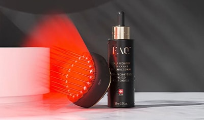 A black, circular device faces outward with a ring of red lights shining from it. It leans against a black serum bottle with gold details.