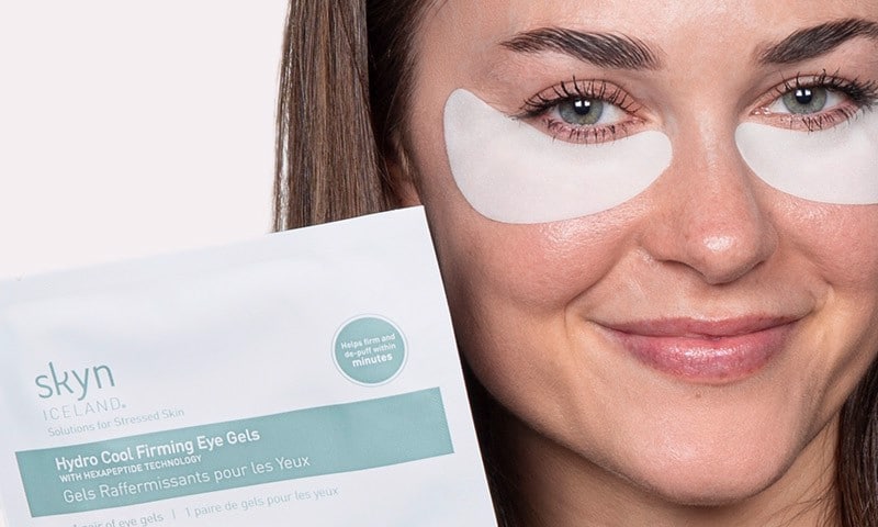 Close-up of person wearing under-eye gel patches holding up the skyn ICELAND Hydro Cool Firming Eye Gels package