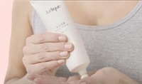 A woman with fair skin, nude colored nails and a grey tank top dispenses Jurlique Rose Hand Cream into her hand.