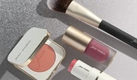 A powder blush in a compact, a cream blush in a tube, and a liquid blush in a tube lay next to a blush brush.