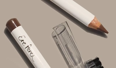 Two Ere Perez Almond Brow Pencils lay on a tan background with a lid and brush.