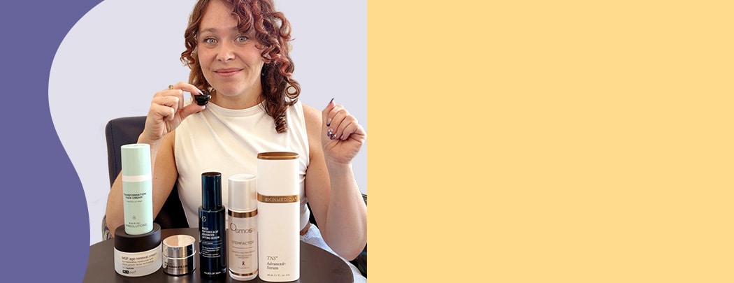 A woman with fair skin and red hair holds a small microphone while sitting behind an array of growth factor skin care products.