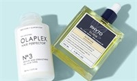 A cylindrical bottle of OLAPLEX No. 3 hair treatment and a rectangular glass bottle of PHYTO hair oil laying down on a light blue background.