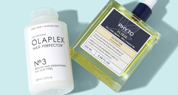 A cylindrical bottle of OLAPLEX No. 3 hair treatment and a rectangular glass bottle of PHYTO hair oil laying down on a light blue background.