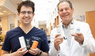 Two dermatologists, one in his 30s with dark hair &one in his 60s with light hair, stand next to one another smiling while holding various sunscreens.