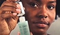 A black woman smiling very softly and pulling the dropper out of a bottle of ELEMIS Pro Collagen Renewal Serum.