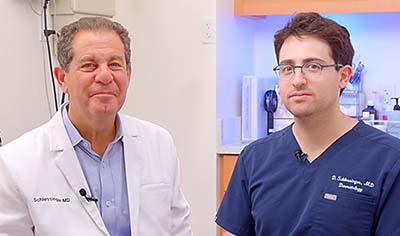 Two dermatologists, one in a white jacket and one in scrubs, sit next to one another in a patient room smiling.