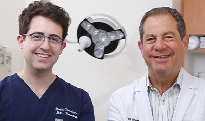 Two dermatologist, one in his 30s with fair skin & dark hair & one in his 60s with light brown hair smile while crossing their arms.