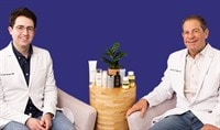 Drs. Joel and Daniel Schlessinger sit next to one another. Between them is a wood table with an array of sunscreens. They are both smiling.