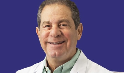Dr. Joel Schlessinger, wearing a light green shirt and a white doctor jacket, smiles at the camera.