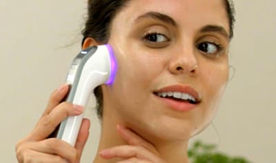 A woman with medium, olive-toned skin holds the Althaea Skin CARA Multi-Modal tool to her right cheek. She is in a bathroom setting.