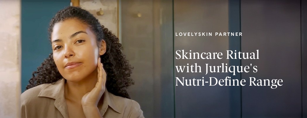 A woman with a medium to deep skin tone and curly brown hair looks into the camera, as if into a mirror, with her hand gently placed below her jaw.