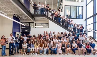 A large group of people who work at LovelySkin are gathered together in the entrance hall of the LovelyGroup headquarters. They all smile.