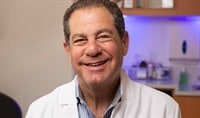 A doctor with light to medium-toned skin and light brown hair wearing a blue shirt and a white doctor's jacket smiles at the camera.