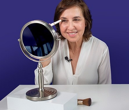 A woman applying an eyeshadow stick to her upper eyelid and smiling in a mirror.