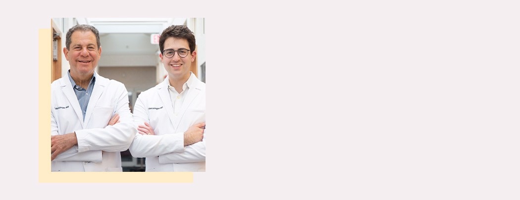 Two male dermatologists, one in his 60s and one in his 30s, stand side by side in white doctor jackets smiling.