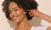 A woman with a medium to deep skin tone and brown curly hair presses a swatch of ISDIN Eryfotona Ageless Tinted Sunscreen SPF 50 to her cheek.