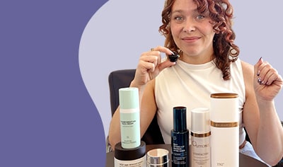 A woman with fair skin and red hair holds a small microphone while sitting behind an array of growth factor skin care products.